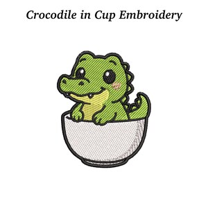 May include: An embroidered design featuring a cartoon crocodile in a white cup. The crocodile is green with a yellow belly and a smiling face. The text "Crocodile in Cup Embroidery" is above the image.