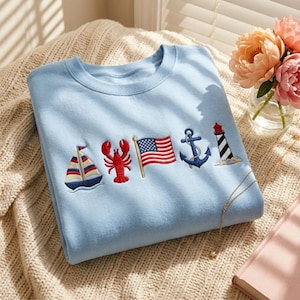 May include: A light blue crewneck sweatshirt featuring a row of embroidered nautical icons across the chest, including a sailboat, a red lobster, an American flag, a blue anchor, and a black and white striped lighthouse with a red top.