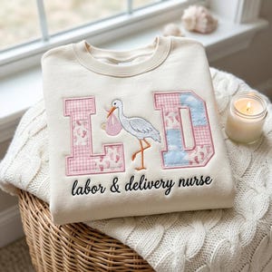 May include: Cream-colored sweatshirt with embroidered design. Features the letters "L D" in pink gingham and blue cloud patterns, with a stork carrying a baby. Text below reads "labor & delivery nurse".