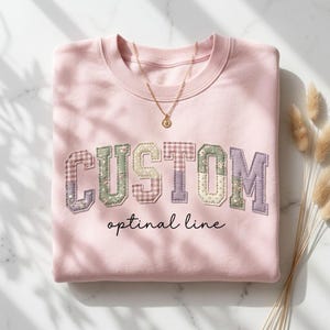 May include: A light pink sweatshirt with the word "CUSTOM" in a patchwork design, featuring floral and gingham patterns. The text "optimal line" is written below the word "CUSTOM". A gold necklace with a pendant is on the sweatshirt.
