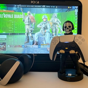 May include: A black and white PlayStation 5 controller stand featuring a skull design, holding a white and black controller. A gaming monitor displays a "Battle Royale Quads" game. A black headset rests in the foreground.