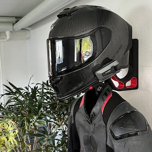 Può includere: Un casco da moto nero in fibra di carbonio con visiera oscurata è montato su un supporto a parete nero e rosso. Il casco ha la scritta "Scorpion" stampata sul lato. Una giacca da moto nera e grigia è visibile sotto.