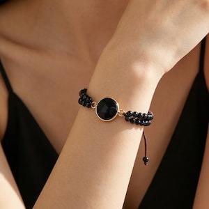 Könnte beinhalten: Schwarzes Perlenarmband mit einem großen, runden, schwarzen Stein in einem goldfarbenen Rahmen. Das Armband befindet sich an einem Handgelenk, der Hintergrund ist ein schwarzes Oberteil.