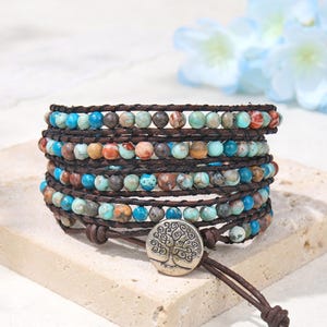 Puede incluir: Una pulsera envolvente de varias vueltas con pequeñas cuentas redondas de jaspe multicolor en tonos turquesa, marrón y beige. Las cuentas están ensartadas en un cordón de cuero marrón oscuro, asegurado con un botón metálico plateado con un árbol.