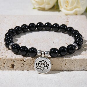 Könnte beinhalten: Schwarzes Perlenarmband mit einem silbernen Lotusblüten-Anhänger. Das Armband hat runde, glänzende schwarze Perlen und silberne Akzente. Der Anhänger ist ein kreisförmiger Anhänger mit einem detaillierten Lotusblüten-Design.