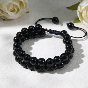 Könnte beinhalten: Ein zweireihiges Armband mit glatten, polierten schwarzen Perlen, die in zwei parallelen Reihen angeordnet sind. Das Armband verfügt über einen verstellbaren schwarzen Kordelverschluss mit zwei kleineren schwarzen Perlen an den Enden.
