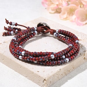 May include: A multi-strand wrap bracelet featuring deep red, speckled glass beads interspersed with smooth, matte silver-toned metallic beads. The bracelet is secured with a brown leather cord and a circular metal button closure at the end.