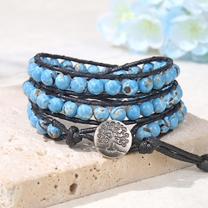Pode incluir: Uma pulseira de várias voltas com três filas de contas redondas azul-brilhante com veios castanho-escuros, enfiadas num cordão preto. Um fecho de botão de metal prateado na extremidade apresenta um design detalhado de uma árvore.