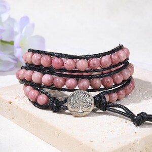 May include: A multi-strand wrap bracelet featuring rows of smooth, round, dusty pink beads strung on black cord. The bracelet is secured with a circular silver-toned metal button closure embossed with a detailed tree of life design.