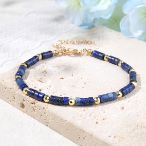 Puede incluir: Una pulsera que presenta una alternancia de cuentas cilíndricas de piedra azul oscuro y pequeñas cuentas metálicas doradas facetadas. La pieza incluye una cadena extensora de metal dorado con un cierre de mosquetón, creando un diseño delicado.