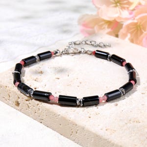 May include: A bracelet featuring alternating black cylindrical beads and small pink round beads, accented with silver-toned metal spacers. The piece includes a silver-toned metal chain extender and a lobster claw clasp for secure closure.