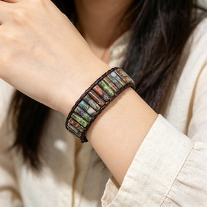 May include: A wrap bracelet featuring rectangular, multi-colored beads in shades of green, brown, and red. The beads are strung on a dark brown leather band. The bracelet is worn on a wrist against a cream-colored shirt.