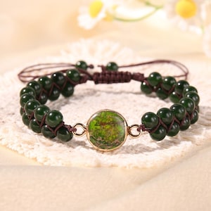 May include: A dark green beaded bracelet with a central circular stone. The bracelet features two rows of dark green beads, connected by brown cord. The central stone is a mottled green color, set in a gold-colored metal frame.