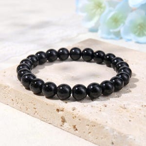 Könnte beinhalten: Ein kreisförmiges Stretch-Armband bestehend aus glatten, polierten schwarzen kugelförmigen Perlen. Die Perlen sind einheitlich groß und in einer durchgehenden Schleife aufgereiht, wobei sie das Licht auf ihren glänzenden Oberflächen reflektieren.