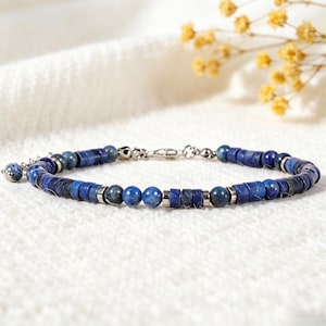 Puede incluir: Una pulsera de cuentas azules con una mezcla de cuentas redondas y rectangulares. La pulsera presenta un cierre plateado y un pequeño dije. Las cuentas son de un tono azul profundo y la pulsera se exhibe sobre una superficie blanca texturizada.