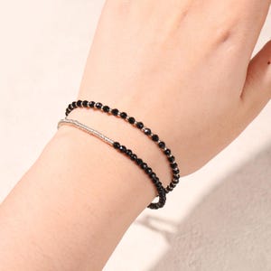 May include: Two beaded bracelets. One bracelet features small, black beads strung together. The other bracelet is silver with small, black beads. The bracelets are worn together on a wrist.