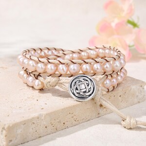 May include: A multi-strand wrap bracelet featuring pale pink pearls strung on light tan cord. The bracelet is secured with a round, silver-toned metal button clasp that displays an intricate, interwoven knot design on its front surface.