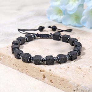 Puede incluir: Una pulsera que presenta cuentas cúbicas de piedra de lava negra alternadas con pequeñas cuentas espaciadoras metálicas de tono plateado. La pieza incluye un cordón negro con un cierre de nudo deslizante ajustable y dos pequeñas cuentas negras.