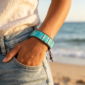 May include: A turquoise and brown bracelet. The bracelet features rectangular turquoise stones strung together with brown leather. The bracelet is worn on a wrist, with a beach in the background.