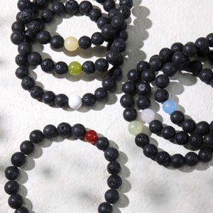 May include: Multiple black lava stone bead bracelets with colourful accent beads. The bracelets feature beads in yellow, green, white, red, and blue. They are displayed on a white surface, highlighting the natural texture and design.