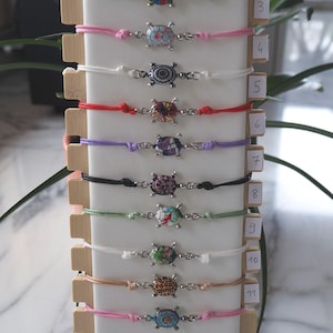 Può includere: Esposizione di undici braccialetti con ciondoli a forma di tartaruga. Ogni braccialetto presenta un cordino di colore diverso e un ciondolo a forma di tartaruga con vari disegni e colori. I braccialetti sono esposti su un supporto di legno con sfondo bianco.