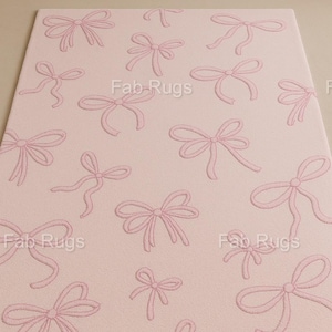 Può includere: Un tappeto rettangolare rosa con un motivo ripetuto di fiocchi rosa chiaro. I fiocchi sono ricamati sulla superficie del tappeto, creando un effetto strutturato. Il tappeto è adatto per una camera da letto o una cameretta.