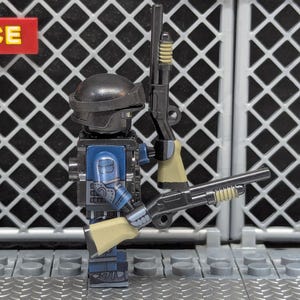 Peut inclure: Une figurine Lego représentant un policier en uniforme bleu et casque noir, tenant deux fusils à pompe jouets. L'arrière-plan présente une grille métallique et une enseigne "SERVICE".
