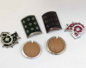 Puede incluir: Colección de seis pines metálicos en forma de escudo. Los pines presentan varios diseños, incluyendo un escudo verde oscuro con tachuelas plateadas, un escudo marrón con tachuelas oscuras y un emblema rojo y plateado. Dos pines son redondos con textura de vetas de madera.