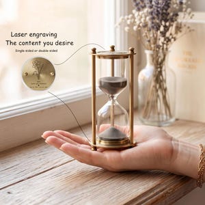 May include: A brass hourglass with clear glass bulbs containing grey sand. The frame features a circular top plate with laser engraving of a flower and text. The text reads: Laser engraving The content you desire Single-sided or double-sided.