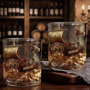 Könnte beinhalten: Zwei klare Trinkgläser mit einem goldenen Wikingerschiff-Design. Das Schiff hat einen Drachenkopf und den Namen "JOHNSON". Die Gläser stehen vor einem Hintergrund aus Weinflaschen und Fässern.