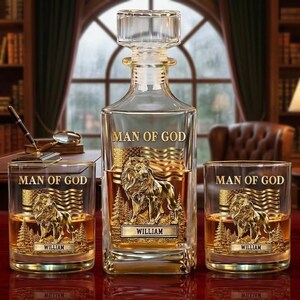 Puede incluir: Un juego de decantador de vidrio transparente con dos vasos a juego. Cada pieza presenta el texto "MAN OF GOD" y un diseño de león dorado con un fondo de bandera. El decantador y los vasos están personalizados con el nombre "WILLIAM".
