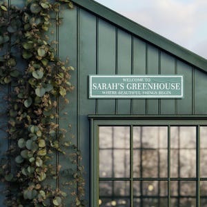 May include: A green wooden sign with white text that reads "Welcome to Sarah's Greenhouse, Where Beautiful Things Begin". The sign is mounted on the side of a green building with a large window.