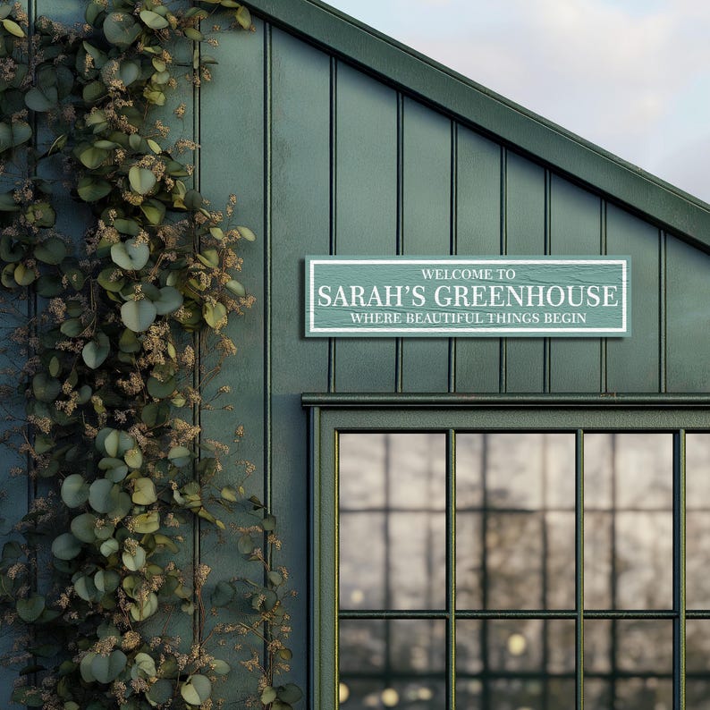 May include: A green wooden sign with white text that reads "Welcome to Sarah's Greenhouse, Where Beautiful Things Begin". The sign is mounted on the side of a green building with a large window.