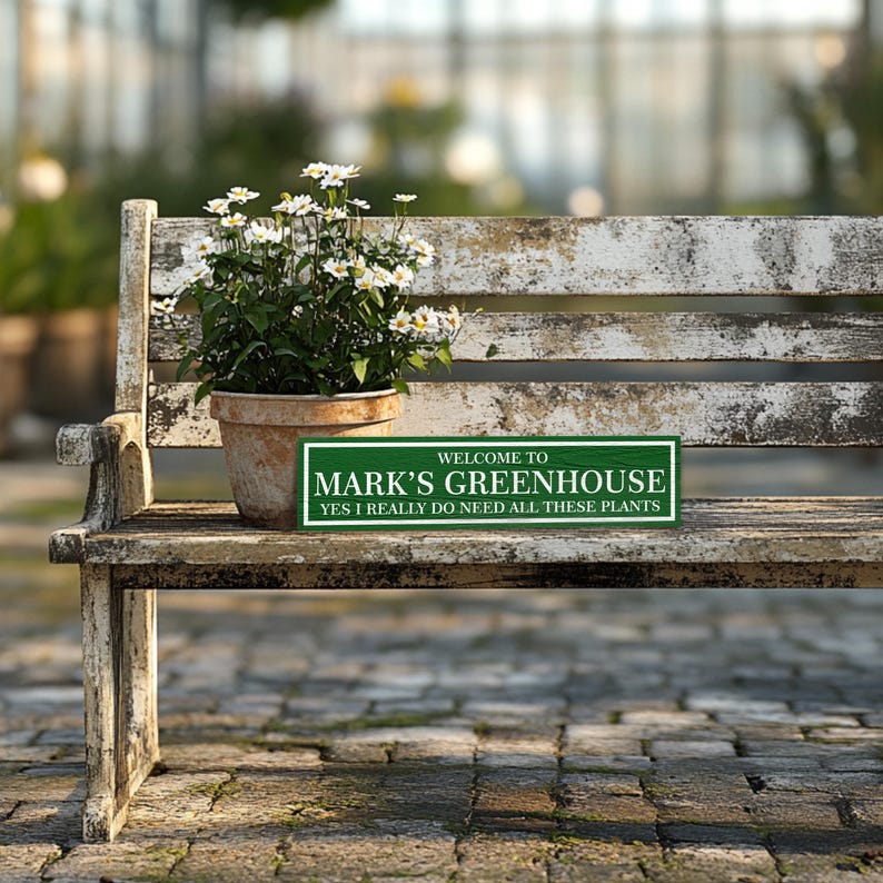 May include: A green wooden sign with white text that reads "Welcome to Mark's Greenhouse, Yes I really do need all these plants". The sign is sitting on a weathered wooden bench with a pot of white flowers.