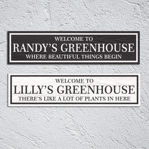 May include: Two black and white wooden signs with white text. The top sign reads "Welcome to Randy's Greenhouse Where Beautiful Things Begin". The bottom sign reads "Welcome to Lilly's Greenhouse There's Like A Lot Of Plants In Here".