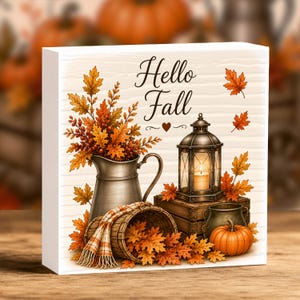 May include: A square white wooden sign featuring the text Hello Fall with a heart icon. The illustration includes a metal pitcher filled with orange and yellow autumn leaves, a lantern with a lit candle, a wooden crate, a basket, and a pumpkin.
