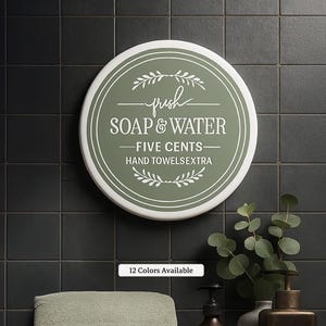 May include: A round, white and green sign with the words "SOAP & WATER" and "FIVE CENTS" in white lettering. The sign also includes the text "HAND TOWELSEXTRA" and decorative leaf accents. The sign is mounted on a dark tile wall.