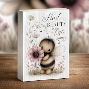 May include: A rectangular wooden block with a watercolor illustration of a bee holding a pink flower. The block has the text "Find BEAUTY in the little things" in script. The background features wildflowers and a wood grain texture.