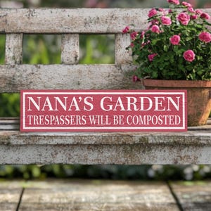 May include: A red wooden sign with white text that reads "NANA'S GARDEN TRESPASSERS WILL BE COMPOSTED". The sign is sitting on a white wooden bench.