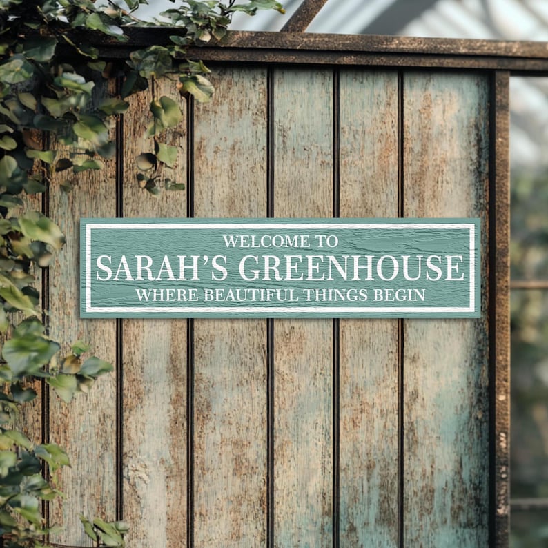 May include: A light blue wooden sign with white text that reads "Welcome to Sarah's Greenhouse Where Beautiful Things Begin". The sign is hanging on a weathered wooden fence.