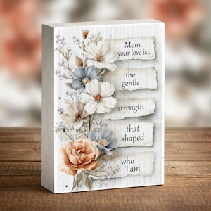 May include: A rectangular wooden block with a floral design and the text "Mom your love is... the gentle strength that shaped who I am." The flowers are white, blue, and peach.