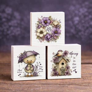 May include: Three white wooden blocks with spring-themed illustrations. One block features a floral wreath with "hello spring" text. Another shows a chick under an umbrella with "April showers bring May flowers." The third block has a birdhouse and the text "Spring is in the air!"