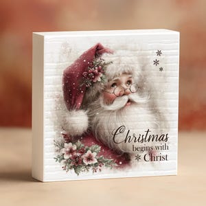 May include: A white wooden block features a watercolor illustration of Santa Claus in a red hat and coat, with a white beard and glasses. The text "Christmas begins with Christ" is written in script.