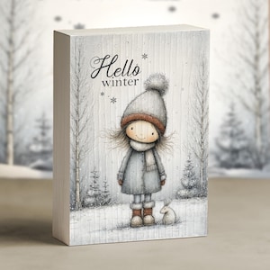 May include: A rectangular wooden block featuring a winter scene illustration. The artwork depicts a girl in a gray coat, hat, and scarf, with a small rabbit. The text "Hello winter" is written in script. The background shows a snowy forest.