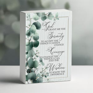 May include: A white wooden block featuring the Serenity Prayer. The block has a framed design with the prayer's text in elegant script. Eucalyptus leaves and branches in shades of green and white adorn the left side, adding a natural touch.