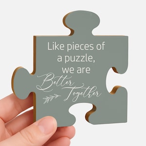 May include: A gray puzzle piece with the words 'Like pieces of a puzzle, we are Better Together' in white script. The puzzle piece is held by a hand and has a light wood grain edge.