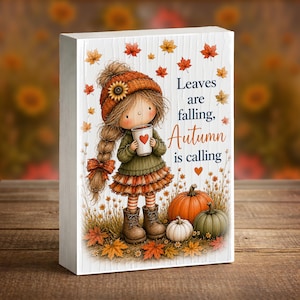 May include: A rectangular white wooden sign featuring a cartoon illustration of a child in an orange knit hat and green sweater holding a cup with a heart. The text reads Leaves are falling, Autumn is calling, surrounded by pumpkins and leaves.