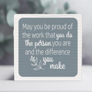 May include: A white wooden block with a gray square featuring the text "May you be proud of the work that you do, the person you are and the difference you make." A floral design is at the bottom.