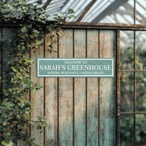 May include: A teal wooden sign with white text that reads "Welcome to Sarah's Greenhouse, Where Beautiful Things Begin".