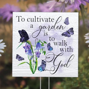 May include: A white wooden garden stake with the text "To cultivate a garden is to walk with God". The stake features a watercolor design of purple flowers and blue butterflies.